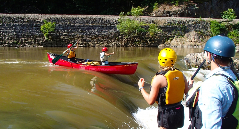 Two people wearing safety gear navigate a canoe over a short waterfall. Two more people stand on the shore, watching them.
